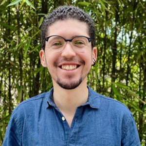 Foto do professor Lucas Fogaça, possui pele clara, olhos e cabelos castanho, lábios finos, usa cavanhaque e óculos com armação escura. Veste uma camisa azul e ao fundo possui vegetação.