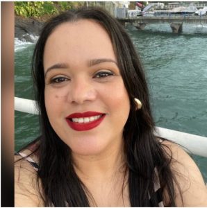 Foto da Professora Patrícia Silva que possui pele clara, cabelos e olhos escuros, usa brinco na orelha esquerda e batom vermelho. Ao fundo, uma passagem de água e ponte.