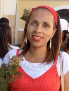 Fotografia. Mulher negra de cabelos lisos e castanhos na altura dos ombros. Ela usa uma faixa vermelha na cabeça, brincos dourados, batom vermelho e usa uma blusa branca sob vestido vermelho. Ela está ao lado de uma planta e sorri. Ao fundo, há pessoas e uma parede clara com detalhes em arco.