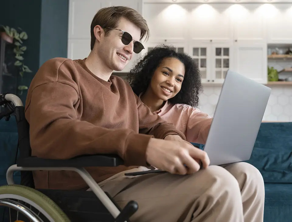 A imagem mostra duas pessoas sentadas juntas em um sofá. A pessoa à esquerda é um homem e está em uma cadeira de rodas e veste uma blusa marrom. Ele está sorrindo e segurando um dispositivo laptop. A pessoa à direita é uma mulher. Ela tem cabelo cacheado e veste uma blusa rosa clara. Ela também está sorrindo e olhando para o dispositivo. Ao fundo, há uma parede com um padrão hexagonal e um móvel branco com portas de vidro.