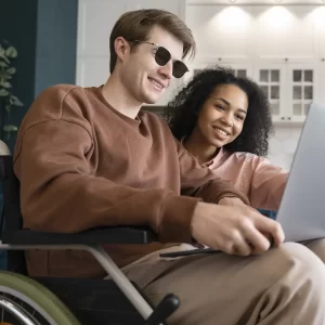 A imagem mostra duas pessoas sentadas juntas em um sofá. A pessoa à esquerda é um homem e está em uma cadeira de rodas e veste uma blusa marrom. Ele está sorrindo e segurando um dispositivo laptop. A pessoa à direita é uma mulher. Ela tem cabelo cacheado e veste uma blusa rosa clara. Ela também está sorrindo e olhando para o dispositivo. Ao fundo, há uma parede com um padrão hexagonal e um móvel branco com portas de vidro.